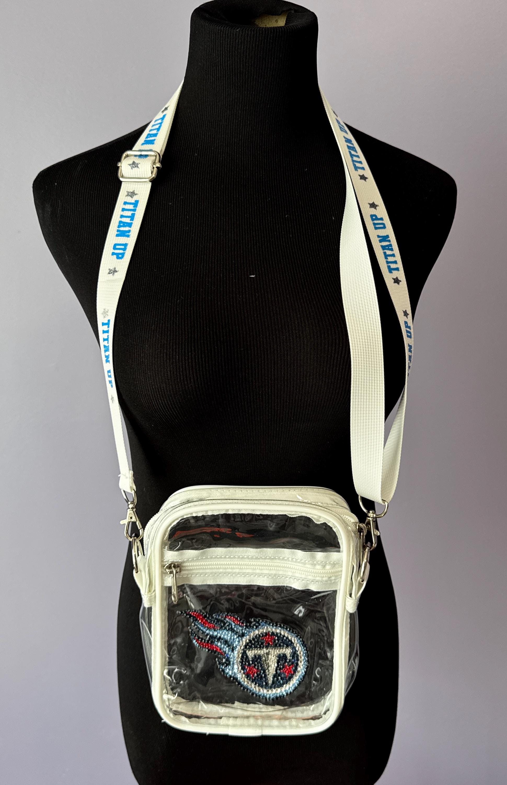 May include: A clear, rectangular crossbody bag with a white trim and strap. The bag features a Tennessee Titans logo on the front. The white strap has &#39;TITAN UP&#39; printed in blue. The bag is displayed on a black mannequin against a gray background.